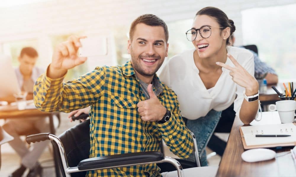 Man in wheelchair taking a selfie with a co-worker at his desk.